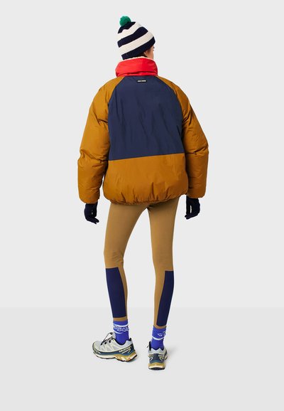 Giubbotto a vento marrone, blu navy e rosso con vestibilità rilassata; abbinato a leggings beige con dettagli blu navy e scarpe sportive. Berretto a righe sulla testa.