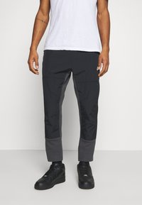 Man in zwarte taps toelopende broek met versterkte knieën en enkelpanelen, een wit T-shirt en zwarte sneakers, staande op een witte vloer.