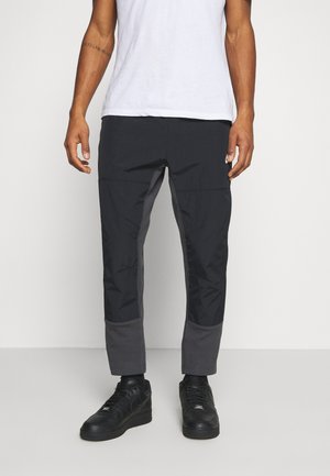 Mann in schwarzen, angepassten Hosen mit verstärkten Kniefeldern und Knöcheln, weißem T-Shirt und schwarzen Sneakern, der auf einem weißen Boden steht.