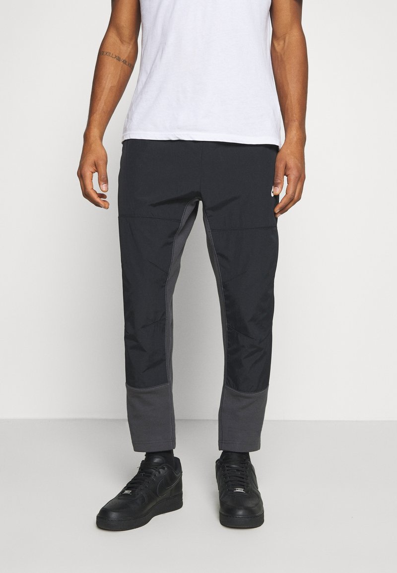 Man in zwarte taps toelopende broek met versterkte knieën en enkelpanelen, een wit T-shirt en zwarte sneakers, staande op een witte vloer.