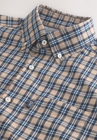 Chemise à carreaux marron, bleu et crème avec un col boutonné, ornée de boutons blancs et d'un petit logo sur le devant. Matière en mélange de coton.