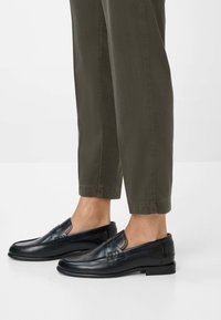 Schwarze Lederloafers mit glatter Oberfläche und dekorativem Stitching, getragen mit olivgrünen, cropped Hosen, die ein minimalistisches Design zeigen.