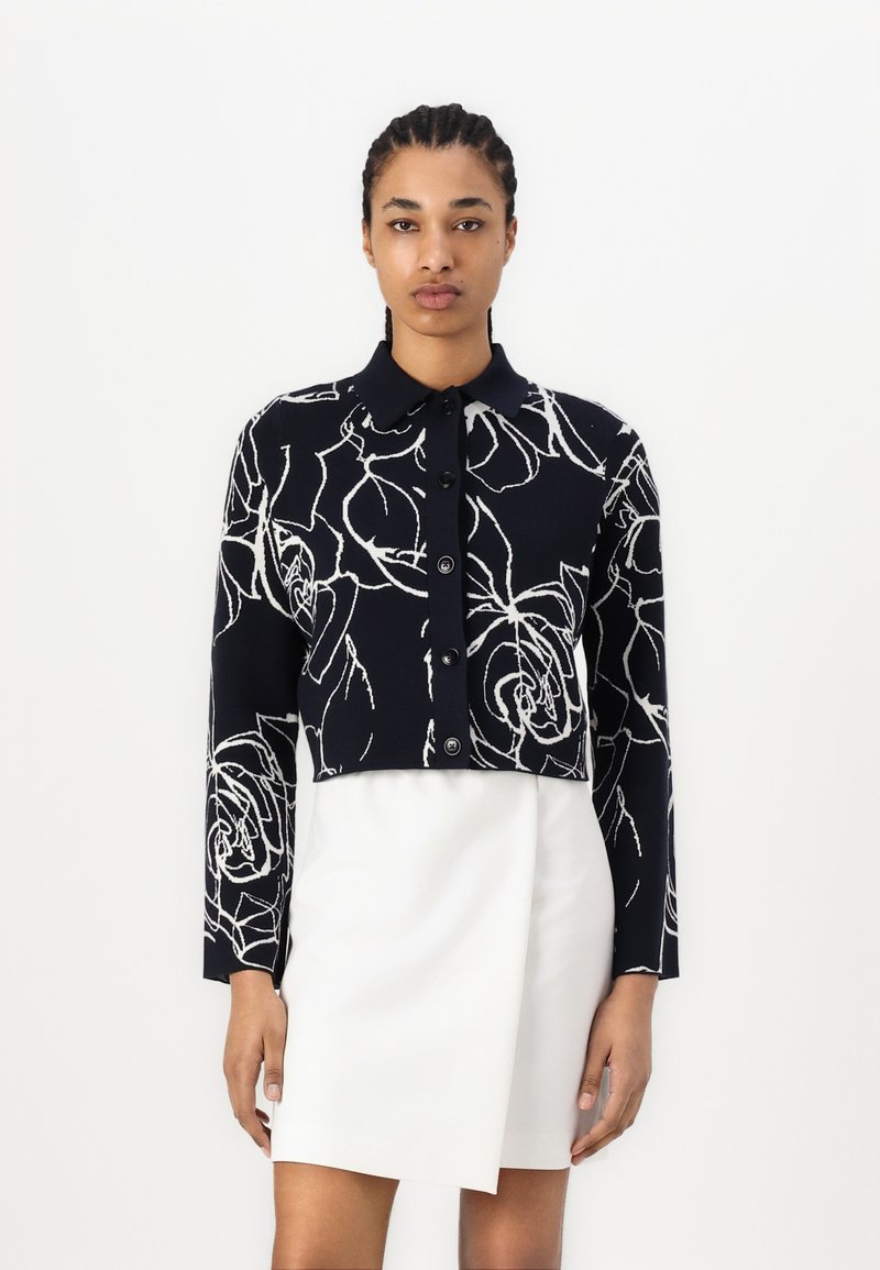 Κοντομάνικο ναυτικό πουλόβερ με λευκό σχέδιο τριαντάφυλλου, μακρύ μανίκι και κουμπιά μπροστά. Φορεμένο πάνω από μια λευκή φούστα, με καθαρή σχεδίαση και λεία υφή.