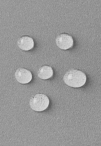 Klare Wassertropfen unterschiedlicher Größen auf einer strukturierten grauen Oberfläche, die runde und ovale Formen mit glatten Kanten und subtilen Reflexionen zeigen.