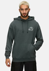 Sudadera con capucha de color gris oscuro, hecha de una tela suave, que cuenta con un bolsillo tipo canguro en la parte frontal y un logo de un casco de fútbol americano en el lado izquierdo del pecho.