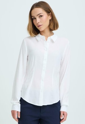 Mujer con cabello hasta los hombros, vestida con una blusa blanca abotonada y pantalones azul marino, de pie frente a un fondo claro.