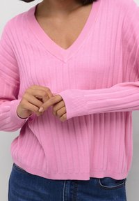 Pull en tricot rose clair avec texture côtelée, col en V et manches longues. L'ourlet est légèrement ample, complété par des poignets retroussés.