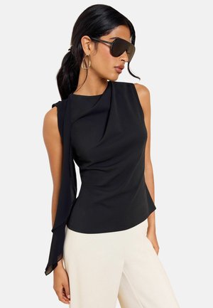Femme aux longs cheveux foncés portant de grandes lunettes de soleil noires, des boucles d’oreilles géométriques dorées, un haut noir sans manches avec un détail drapé et un pantalon blanc.