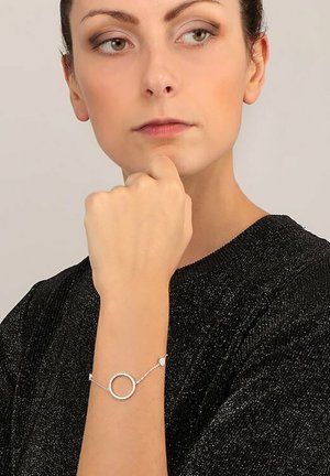 Bracciale d'argento con pendente rotondo e piccole pietre preziose, posizionato su una superficie nera texturizzata. Il design circolare mette in risalto l'eleganza.