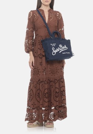 Mujer con un vestido largo marrón con ojales y sandalias, llevando un bolso tote azul marino Saint Barth con texto blanco y bordes deshilachados.
