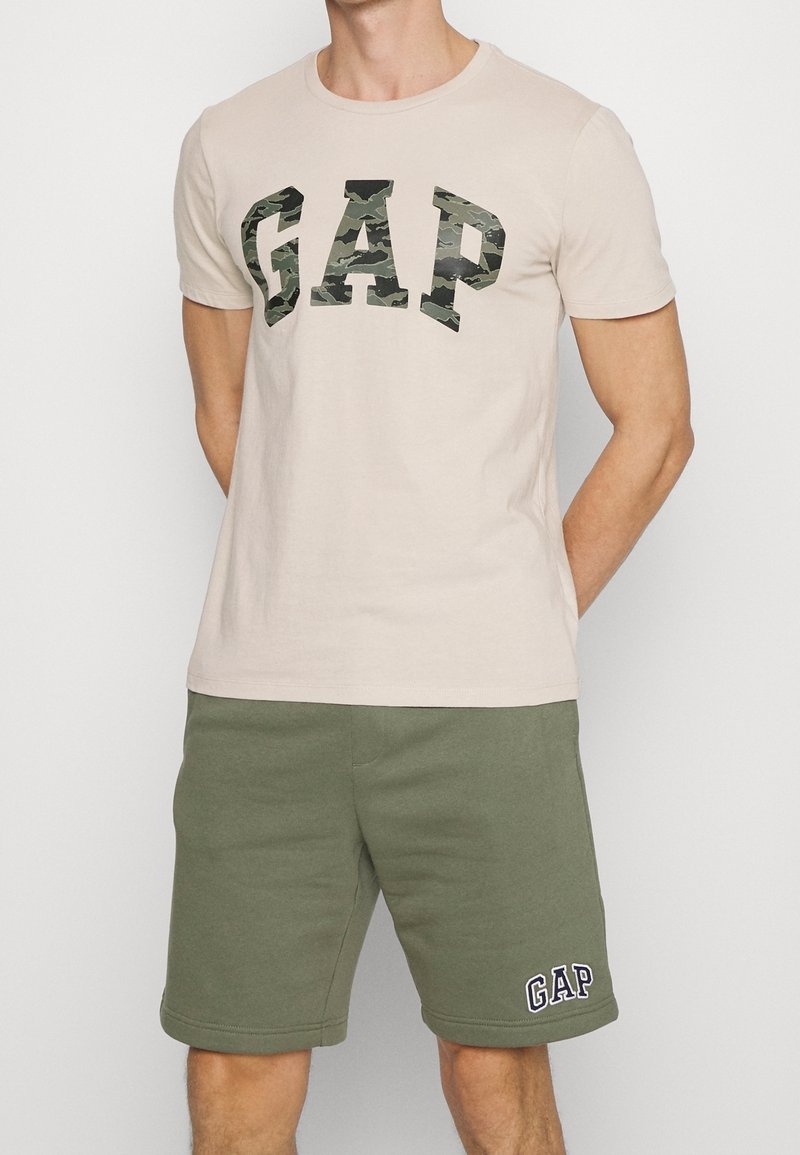 Camiseta de algodón beige con letras de camuflaje negro y verde que forman la palabra "GAP", combinada con unos pantalones cortos de forro polar verde oliva con un logo bordado.