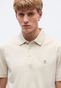 Béžové polo tričko s klasickým golierom, tromi gombíkmi a malým kruhovým logom na ľavej strane hrude. Hladká textúra a priliehavý strih.