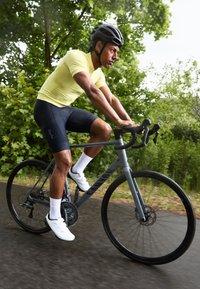 Ein Radfahrer in einem gelben Fahrradtrikot und schwarzen Shorts fährt auf einem grauen Canyon-Bike mit schwarzen Rädern auf einem von Bäumen gesäumten Weg.