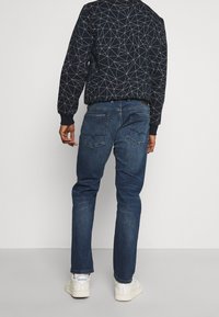 Dark blue denim jeans with a slim fit, featuring two back pockets and minimal stitching detail. Paired with a patterned black bomber jacket.