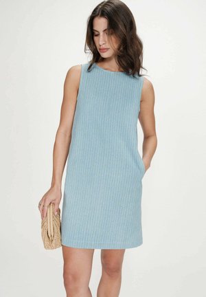 Femme portant une robe sans manches rayée bleu clair, tenant une pochette tissée dans sa main gauche, la main droite dans sa poche.