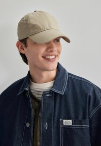 Khaki baseballkorkka, jolla on rakenteellinen muoto. Yhdistettynä tummansiniseen farkkutakkiin, joka sisältää valkoisia ompeluksia ja taskumerkin.