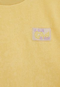 Žuti tkanina s teksturom i malim komadom s prikazom kriški limuna i tekstom "Le Club De Soleil" ljubičastim slovima.