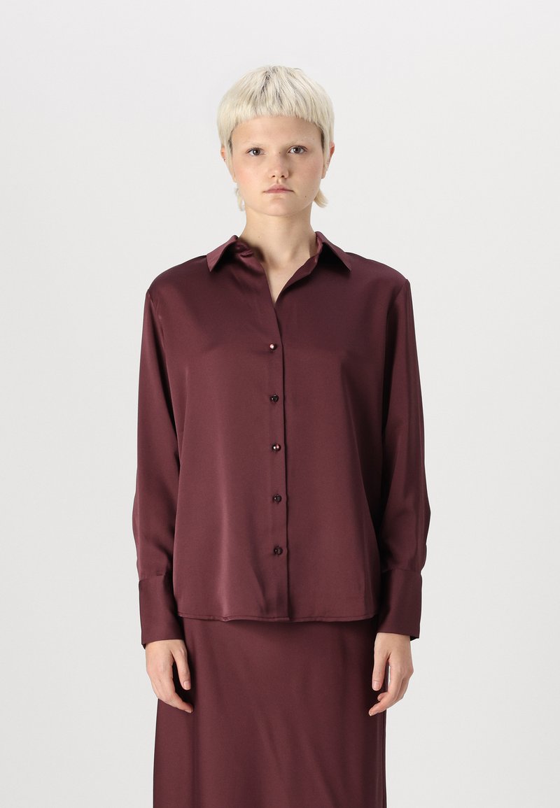 Bordová saténová košeľa s gombíkmi, golierom, dlhými rukávmi a predným zapínaním. Hladká textúra, mierne uvoľnený strih a viditeľná zodpovedajúca sukňa.