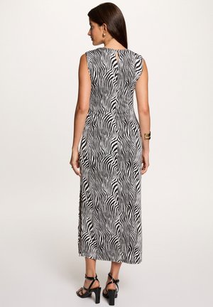 Vrouw staand, gekleed in een mouwloze midi-jurk met zwart-witte zebraprint en zwarte sandalen met hoge hakken, met een gouden armband aan haar rechterpols.