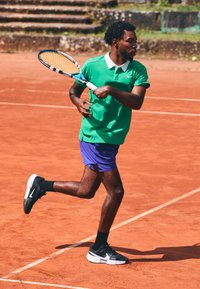 Tennisist, kes kannab rohelist polo särki ja lillat lühikeste pükse, hoiab kätetennisereket. Mustad ja valged tossud, jookseb saviväljakul.