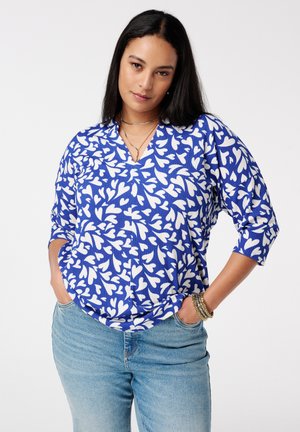 Vrouw met lang, donker haar die een blauw-wit topje met hartjespatroon en lichtblauwe spijkerbroek draagt, staand met handen in de zakken.