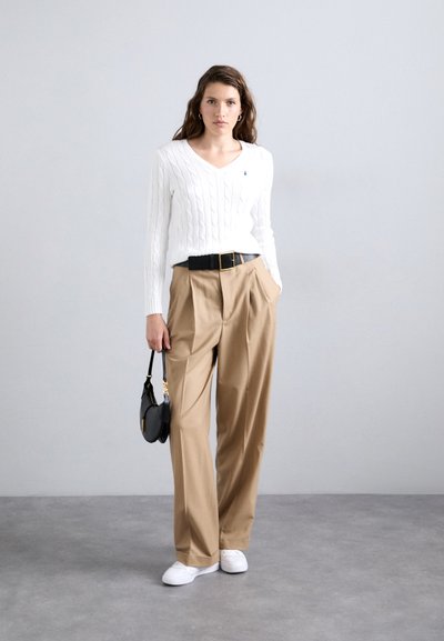Un pull en maille blanche avec décolleté en V, un pantalon plissé beige, une ceinture noire et un sac à main noir. Des baskets blanches complètent le look.