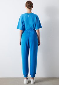 Blue short-sleeve top with crew neck and back logo, paired with matching jogger pants featuring elastic cuffs and waist. White sneakers.