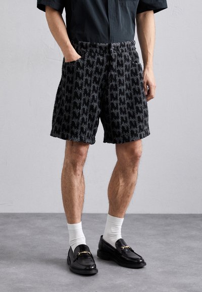 Shorts noirs texturés avec un motif en relief. Présentent une taille élastique et des poches latérales. Portés avec des mocassins noirs et des chaussettes blanches.