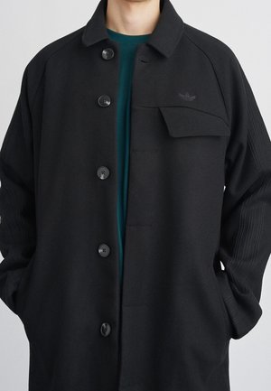 Homme portant un manteau noir boutonné avec une poche poitrine à rabat et un logo discret, les mains dans les poches latérales, sur une chemise turquoise.