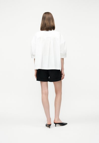 Femme debout, portant un chemisier blanc ample, un short noir et des mules à talons noires, vue de dos sur un fond clair uni.