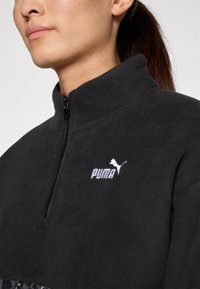 Black fleece pullover with high collar, half zip, and Puma logo in white. Features a smooth texture and contrasting lower panel with a pattern.