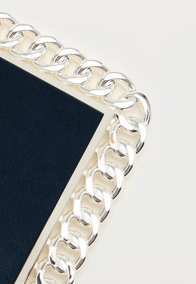 Κλείσιμο εικόνας μιας ορθογώνιας ασημένιας αλυσίδας που περιβάλλει επιφάνεια με σκούρο μπλε ναυτικό χρώμα και υφή, σε λευκό φόντο.