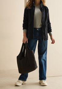 Black lightweight jacket with zip, gray sweater, blue jeans, and white sneakers. Holding a dark quilted bag with shoulder straps.