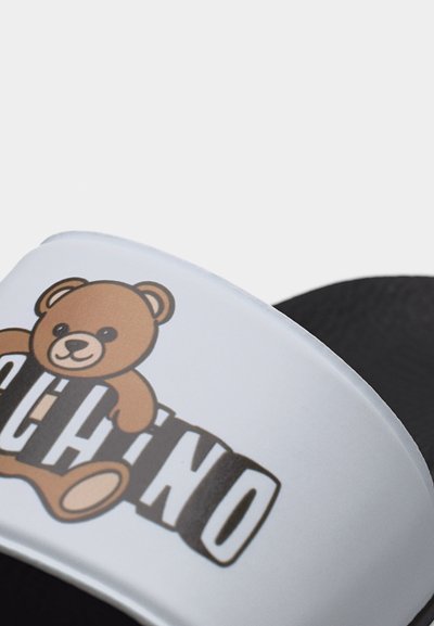 Λευκό λουρί σανδάλι με καφέ αρκουδάκι και το κείμενο "MOSCHINO" σε μαύρο και λευκό σε μαύρη σόλα.