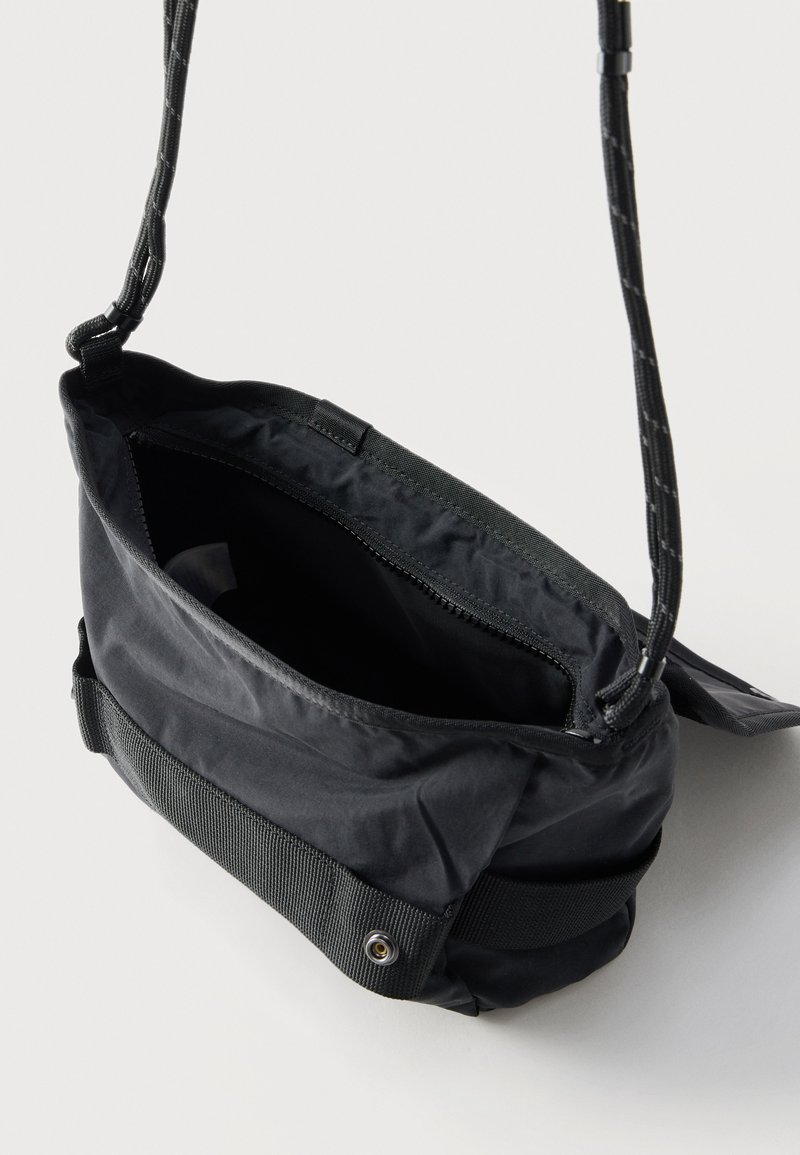 Zwarte crossbodytas met verstelbare riem, bovenste ritssluiting en vooraan drukknopdetail, afgebeeld op een effen lichte achtergrond.