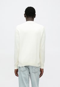 Camisola creme com desenho em malha texturizada, decote redondo e punhos e bainha canelados. Usada por cima de jeans azul claro, exibindo um ajuste descontraído.