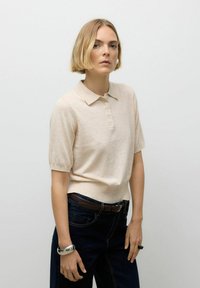 Helle beigefarbene Kurzarm-Polohemd mit Kragen und fünf Knöpfen, kombiniert mit dunkelblauen hochtaillierten Jeans und einem silbernen Armband.