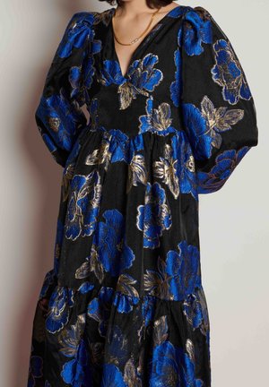 Femme portant une robe noire avec des motifs floraux bleus et dorés, des manches bouffantes et un collier en chaîne dorée.