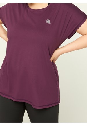 Persoon draagt een loszittend paars shirt met korte mouwen en een wit abstract logo op de borst, gecombineerd met een zwarte broek, handen in de zij.