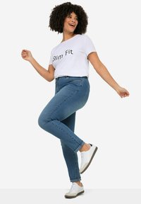 Úzky biely tričko s čiernym textom, modré džínsy a biele tenisky so šedými akcentmi. Pozícia je uvoľnená s prekročenou nohou.