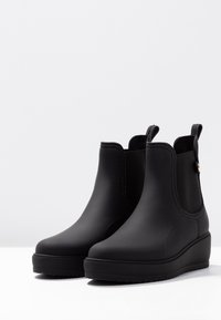 Bottes de cheville en caoutchouc noir avec une finition mate, des panneaux élastiques et une semelle en wedge texturée, dotées de languettes en boucle pour un enfilage facile.