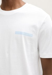 Balta kokvilnas T-krekls ar apaļu apkakli, uz kura kreisajā pusē redzams gaiši zils "DENIM" grafiskais attēls. Īsas piedurknes un gluda tekstūra.