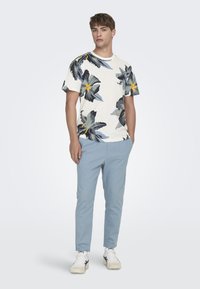 Kortsleeve kremfarget t-skjorte med et fargerikt blomstermønster, kombinert med lyseblå smale bukser og hvite joggesko med svarte detaljer.