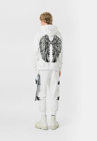 Sudadera blanca con diseño de alas de lentejuelas negras y plateadas en la parte posterior. El pantalón de chándal blanco a juego presenta acentos de alas negras. Botas de combate blancas.
