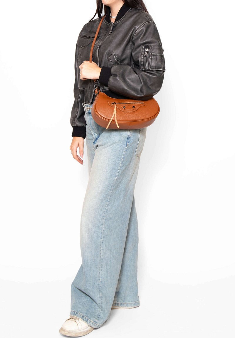 Borsa a tracolla in pelle marrone con forma arrotondata, superficie testurizzata e dettaglio decorativo con lacci. Abbinata a una giacca bomber nera e jeans chiari.