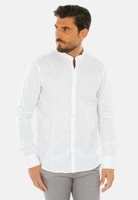 Camicia bianca a maniche lunghe con colletto rigido, chiusura con bottoni e design aderente. Tessuto liscio senza motivi o dettagli visibili.