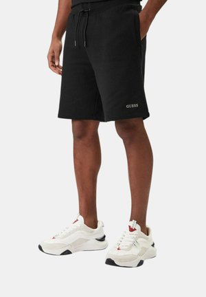 Uomo che indossa pantaloncini neri con coulisse e logo Guess e sneaker bianche, in piedi davanti a uno sfondo semplice.
