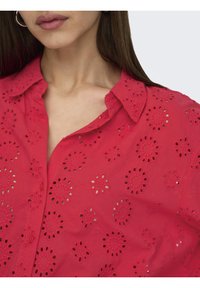 Femme portant une chemise rouge à boutons avec un motif floral en œillets et un col, montrant une partie de son cou et de ses lèvres.