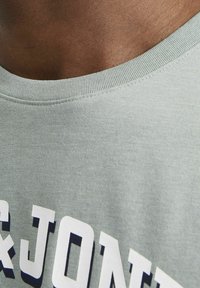 Lys grå bomuld t-shirt med ribbet halsudskæring. Stort hvidt trykt tekst foran med subtil skygge. Glat, let tekstureret stof.