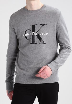Mężczyzna w szarej bluzie Calvin Klein Jeans i czarnych spodniach, stojący na tle jasnego, jednolitego tła.